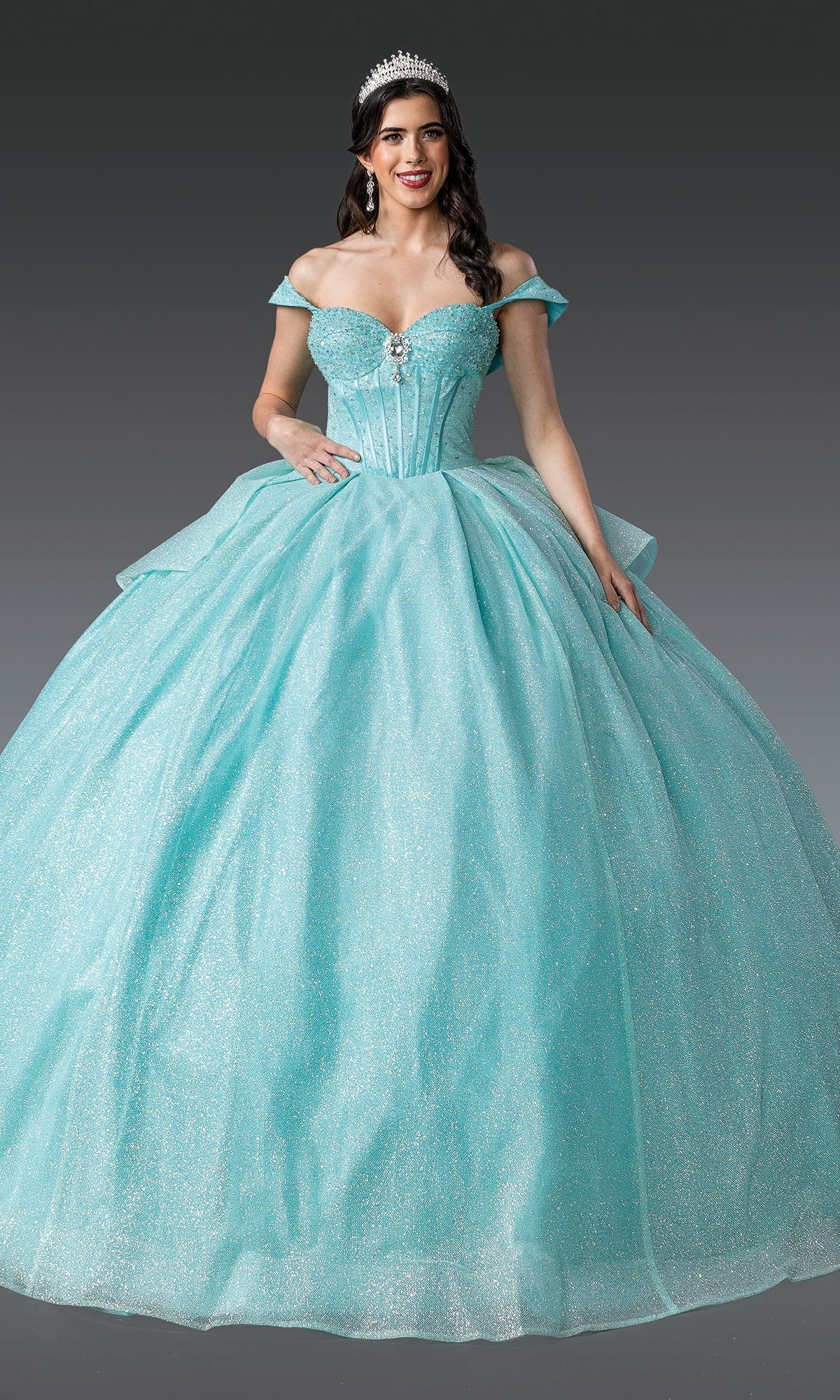 A woman in the Quinceanera Dress 1937 By Dancing Queen, a sparkling light blue ball gown with off-the-shoulder straps and a tiara, stands against a plain gray background.